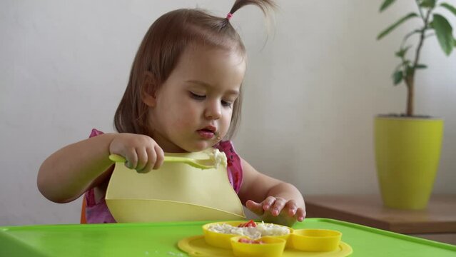 Close up of lovely baby girl eating have breakfast eggs vegetables sausage by herself at home. Adorable possitive happy toddler child eats with dirty face at table indoor. Food, nutrition healthy life