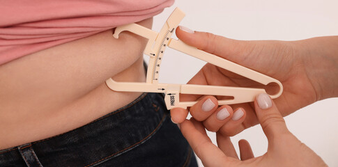 Nutritionist measuring overweight woman's body fat layer with caliper on white background, closeup....