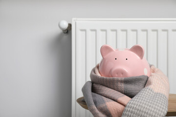 Piggy bank wrapped in scarf on table near heating radiator, space for text