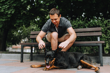 A man sits on a bench in the park, plays and caresses his beloved Rottweiler dog lying on the tile....