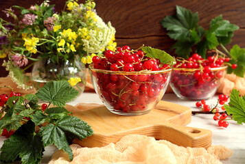 Red currants in a glass cup on a wooden kitchen board against a background of a bouquet of summer flowers, currant branches, wooden background, close-up