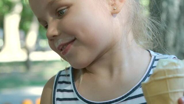 Smiling little girl eats ice cream looks at the camera and flirts. Close-up portrait of blonde girl sitting on park bench and eating ice cream and smiles. Slow motion, Camera moves around