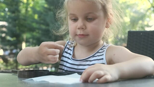 Little girl tastes salt. Close-up of blonde girl takes salt from napkin with her finger and tastes it while sitting in street cafe on the park. Slow motion.