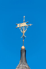 An FS weather vane is attached to the top of a peaked roof. The weathervane has an arrow and a filigreed base. A blue sky is behind the weathervane