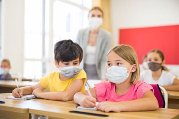 Focused preteen pupils in protective face masks studying in classroom with female teacher. Necessary precautions in coronavirus pandemic