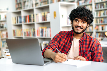 Photo of a handsome positive student or freelancer, arabian or indian nationality with curly hair,...
