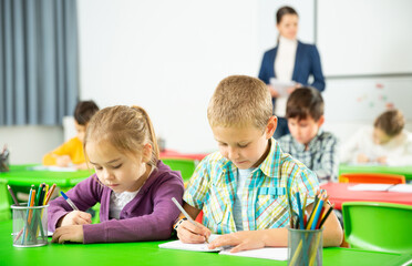 Fototapeta premium Assiduous school kids with pens and notebooks studying in classroom with female teacher