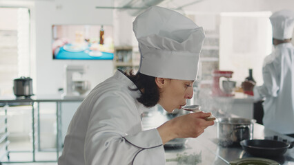 Female chef tasting liquid sauce on stove to make cuisine dish, cooking culinary recipe with fresh ingredients. Woman with uniform working in restaurant kitchen to create professional menu. Close up.