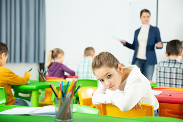 Fototapeta premium Portrait of tired schoolgirl sitting at pupils desk at lesson in primary school