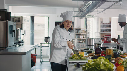 Portrait of female cook cutting green celery in gourmet kitchen, using cutting board and knife to slice coleslaw salad. Woman chef with uniform preparing ingredients for food recipe.
