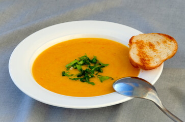 Cream of pumpkin soup with herbs and a slice of toasted bread in a white plate on a gray background. Close-up