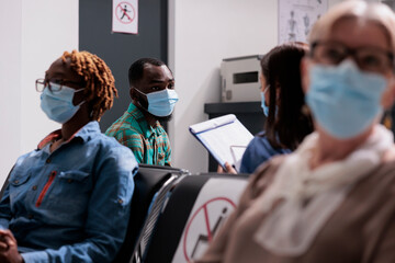 African american patient with face mask talking to nurse, sitting in waiting area at hospital reception lobby. Man doing healthcare consultation with medical worker at facility.