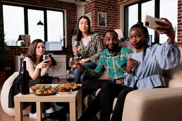 Multiethnic group of friends taking selfie photo together while at wine party in living room. Happy smiling adults at home enjoying time together while drinking alcoholic beverages and taking pictures