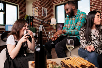 Close diverse friends having fun at wine party while bonding in living room at home. Cheerful people celebrating birthday event with alcoholic beverage and snacks while enjoying time together.