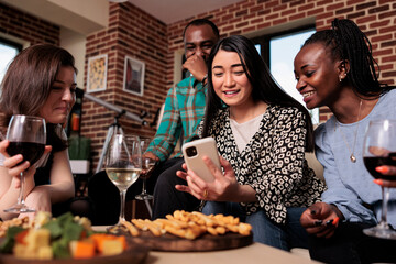 Happy young people having a glass of wine and eating snacks while enjoying time together. Diverse group of friends at wine party celebrating birthday while watching funny clips on smartphone