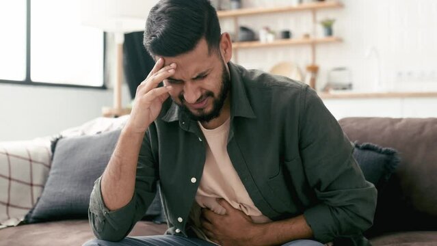 Sad exhausted guy of indian and arabian ethnicity, sits on a sofa in the living room, holds his hands on his stomach, grimaces from pain in his stomach, suffers from poisoning, spasm, stomach problems