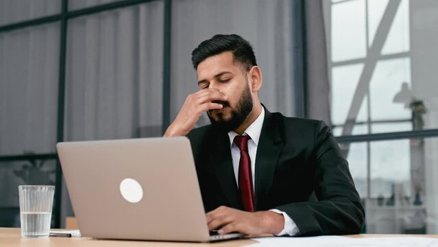 Tired exhausted indian or arabian businessman in a suit, sit at workplace in office, rubbing his nose bridge, feelings fatigue after long time work with laptop, overworked, need rest, coffee break