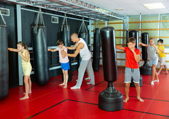 Group of kids boxing with punch bags during their group training. Trainer standing nearby and correcting them. - obrazy, fototapety, plakaty