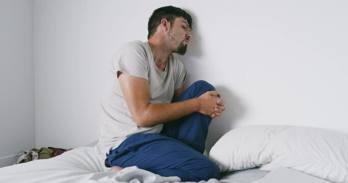 A paranoid creepy man sitting on a bed alone with weird behavior. A young male with mental health issues or suffering from anxiety disorder inside a room. A disturbed and fearful alcoholic guy