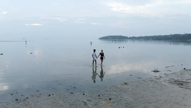 Interracial travelers couple holding hands walking out of shallow sea water on island beach at sunset  - spinning aerial
