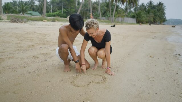 Interracial couple with age differences drawing a heart shape on sandy beach and kissing during a tropical island vacation in Thailand