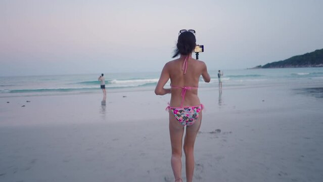 Back view of Thai girl in bikini running on tropical beach during phone live stream at sunset 