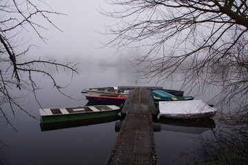 Boote liegen an einem Steg. Es ist Winter und stark nebelig, die Landschaft ist unklar. Von der...