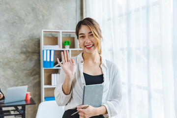 Portrait of friendly asian business woman saying hello, waves her hand and smiles, greets you, hi...