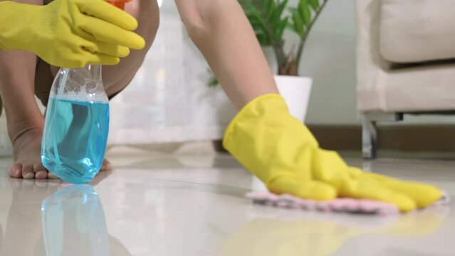 Asian woman wearing yellow rubber glover with cloth rag and detergent spray cleaning floor at home in living room, Female hands wash cleaner, housework and housekeeping concept