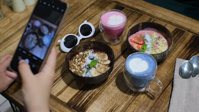 Food blogger taking photos of vegan breakfast with smart phone: blueberry smoothie bowl, strawberry smoothie bowl, pink and blue latte. Slow motion, selective focus