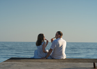 a girl and her dad are sitting on the pier and eating ice cream on a summer evening