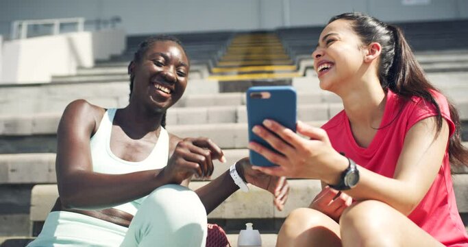 Female athletes laughing while looking at a funny video online together at a stadium. Sporty female friends and teammates enjoying using social media and searching internet at a sports ground
