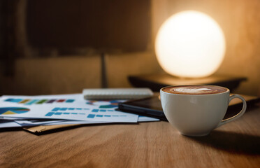 Close up view, Latte coffee in white cup on wooden table with paperwork of chart and tablet computer behind. circle lamp with warm light on background, business concept, retro style and vintage light