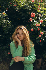 young woman with curly blond hair posing near a blooming rose bush. portrait of a beautiful plus size woman in a green shirt near a bush of flowers in the garden.