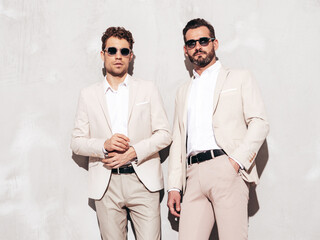 Portrait of two handsome confident stylish hipster lambersexual models. Sexy modern men dressed in white elegant suit. Fashion male posing in studio near grey wall. In sunglasses