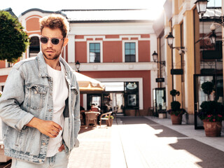 Portrait of handsome confident stylish hipster lambersexual model. Sexy man dressed in jeans jacket. Fashion male posing in the street. In sunglasses in Europe city. Outdoors at sunset