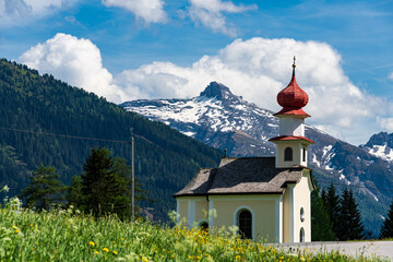 Eine kleine Kapelle auf einer grünen Wiese am Straßenrand einer Landstraße in den Alpen im Sommer