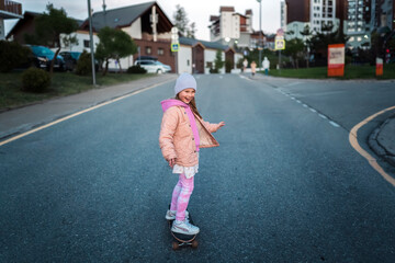 A girl in a pink jacket and hat rides a skateboard on the road