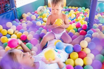 The older sister plays with her younger brother and throws small balls at him while sitting in the playroom