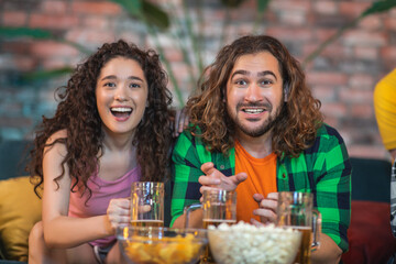 At home in living room attractive couple watching a favourite sport match after their team getting the victory they are hugging each other and are excited lady eating some snacks