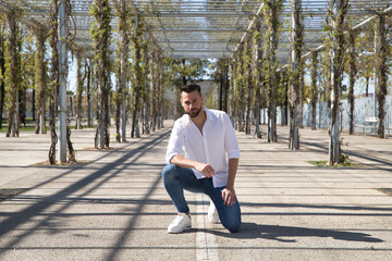 Handsome young man with beard, wearing white shirt and jeans, with one knee on the ground and hands on legs. Concept fashion, beauty, modern, trend, model.