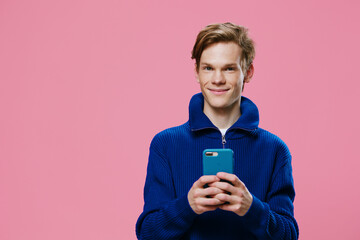 a man stands on a pink background in a blue sweater with a phone in his hands and looks at the camera