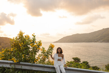 Pretty young caucasian girl in good mood walks in fresh air against backdrop of sea. Brunette wears white top, shirt and pants in summer. Happy day concept