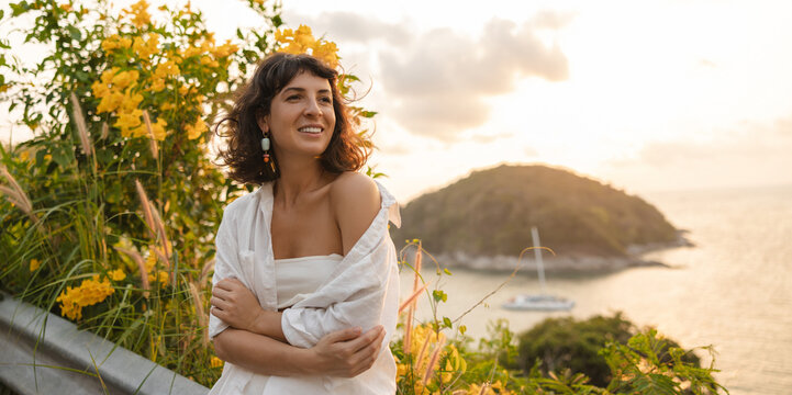 Cute young caucasian girl smiling looking away on background of flower plants and sea. Brunette wears white casual suit. Summer photo session concept
