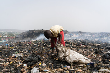 Young boy at a smoky African landfill collecting valuable items for resale, surrounded by clouds of...