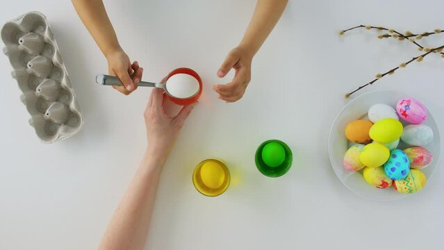easter, holidays and people concept - hands of child and parent dyeing eggs in glass with color on white backhround, top view
