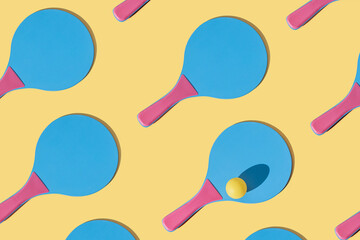 Many blue and pink colourful beach tennis paddle racket with a yellow ping pong ball against yellow background. Flat lay pattern with repetition .Outdoors summer activity, fun relax sport idea.