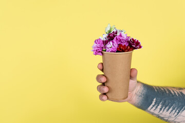 partial view of tattooed man holding disposable cup with natural flowers isolated on yellow.