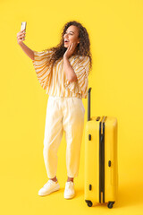 Young African-American woman with suitcase taking selfie on yellow background