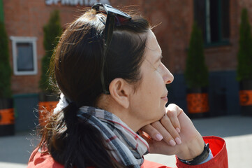 Obraz premium Close-up portrait of a pensive woman looking away, wearing a red jacket, against the backdrop of a city building. Beautiful woman with a pensive expression is resting outdoors in the city.